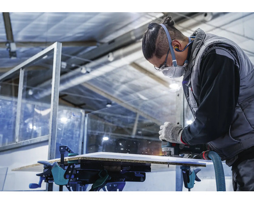 Ein Handwerker fräst eine Platte mit einer Oberfräse, die mit Schraubzwingen auf einer Werkbank befestigt ist.