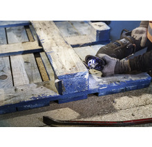 Eine Person sägt eine Holzpalette mit einer Säbelsäge. Ein Brecheisen liegt auf dem Boden.
