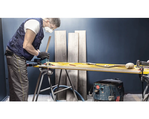 Handwerker schneidet Laminat mit einer Stichsäge auf einer Werkbank. Im Hintergrund stehen Laminatdielen.