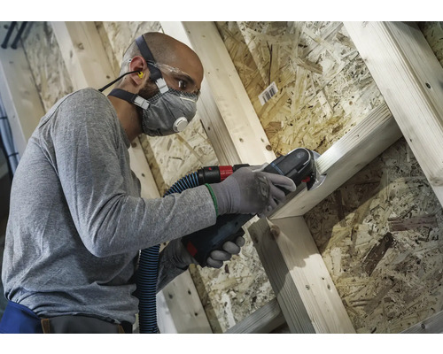 Mann arbeitet mit Multifunktionswerkzeug an Holzkonstruktion mit Staubabsaugung, Atemschutzmaske, Schutzbrille und Ohrstöpseln für Arbeitsschutz