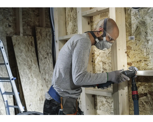 Handwerker mit Atemschutzmaske und Schutzbrille beim Arbeiten mit einem Multifunktionswerkzeug in einem Rohbau