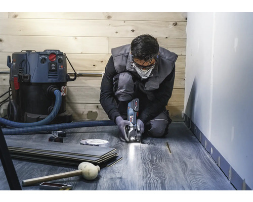 Ein Mann kniet und schneidet Laminat mit einem Multifunktionswerkzeug. Ein Staubsauger, Laminatdielen und Werkzeuge sind ebenfalls sichtbar.