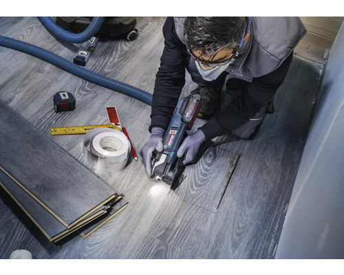 Ein Handwerker schneidet Laminat mit einer Multifunktionswerkzeugmaschine, um einen Ausschnitt für ein Rohr zu schaffen. Werkzeuge wie ein Maßband und ein Bleistift liegen bereit.