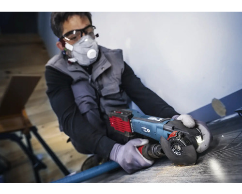 Mann arbeitet mit einem Winkelschleifer auf einem Bodenbelag, persönliche Schutzausrüstung ist vorhanden