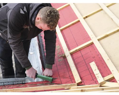 Ein Mann befestigt eine Dachlatte auf einer roten Unterspannbahn mit einem grünen Tacker.