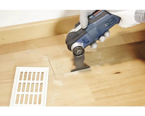 Eine Person schneidet mit einem Multifunktionswerkzeug ein Loch in einen Holzboden neben einem Lüftungsgitter.