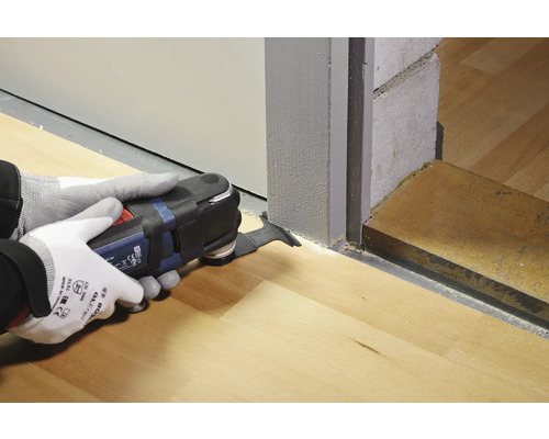 Eine Person verwendet ein Multifunktionswerkzeug, um eine Türzarge auf einem Holzboden zu bearbeiten.