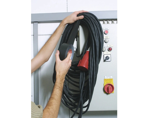 Eine Person befestigt eine schwarze Kabeltrommel mit schwarzem Isolierband an einer Wand.