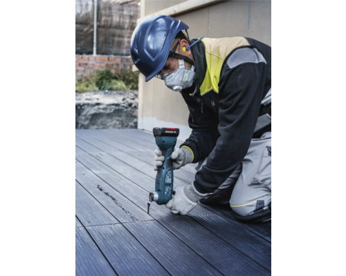 Ein Bauarbeiter mit Schutzhelm und Gesichtsmaske benutzt ein Multifunktionswerkzeug, um eine Nut in eine Terrassendiele zu schneiden.