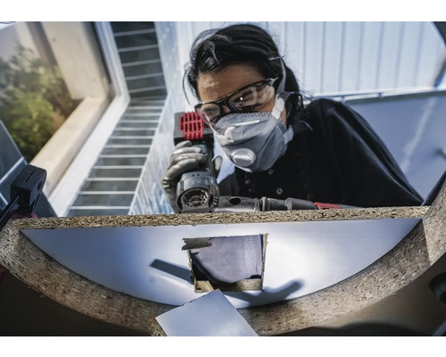 Frau mit Schutzbrille und Staubmaske arbeitet mit einem Multischleifer an einem Werkstück