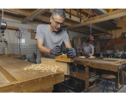 Mann hobelt Holz an einer Werkbank mit Hobel und Handschuhen.