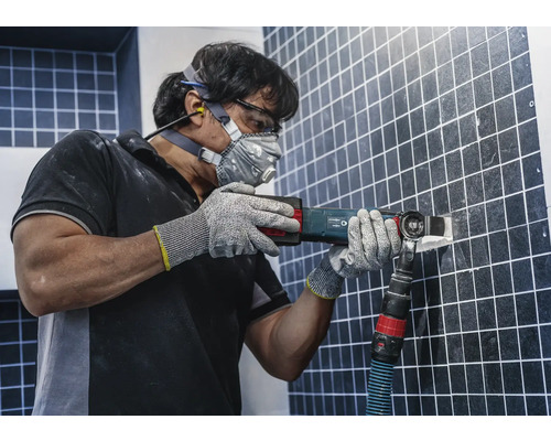 Mann entfernt Fliesen mit einem Multitool, ausgestattet mit Schutzbrille, Gehörschutz, Staubmaske und Handschuhen