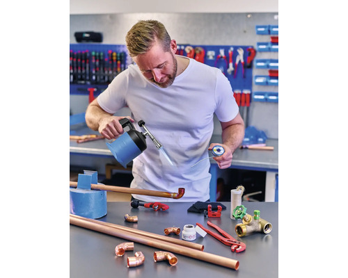 Mann lötet Kupferrohre mit einem Brenner, Lot und verschiedenen Werkzeugen auf einer Werkbank.