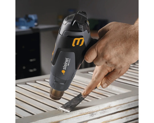 Eine Person entfernt Farbe mit einer Heißluftpistole und einem Spachtel von einem Fensterrahmen.