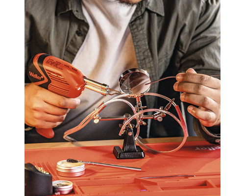 Eine Person lötet mit einer Lötpistole Drähte, die mit einer Halterung mit Lupe fixiert sind. Auf der Arbeitsfläche befinden sich Flussmittel und Reinigungszubehör.