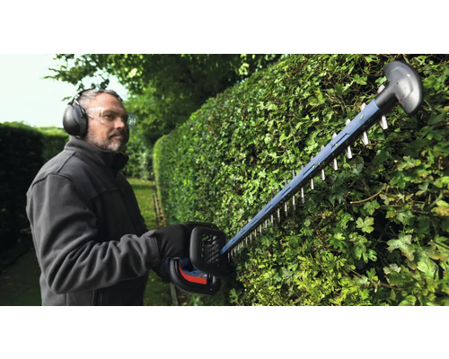 Mann schneidet Hecke mit Heckenschere und trägt dabei Schutzausrüstung.