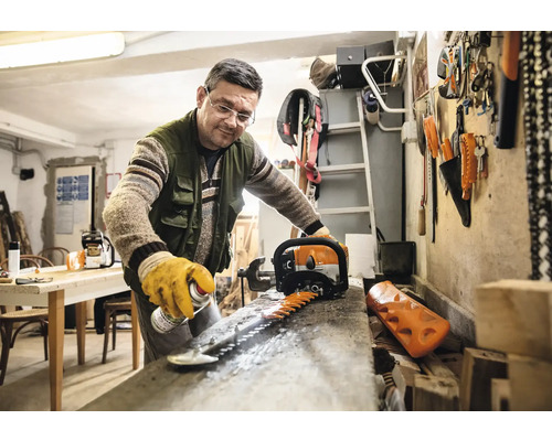 Ein Mann reinigt die Klinge einer Heckenschere mit einem Spray in einer Werkstatt.