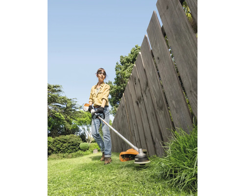 Eine Frau benutzt einen Rasentrimmer im Garten, um Gras entlang eines Holzzauns zu schneiden.