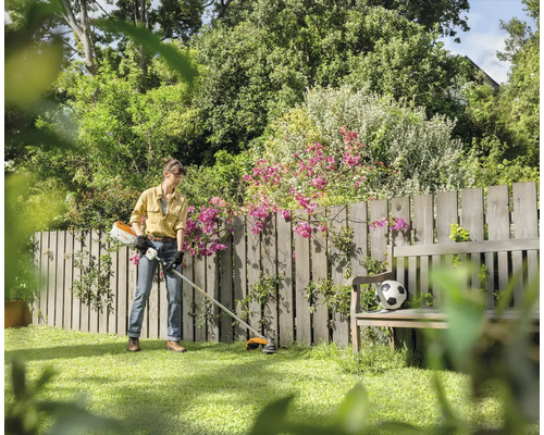 Eine Person verwendet einen Rasentrimmer in einem Garten mit Holzzaun und einer Holzbank.