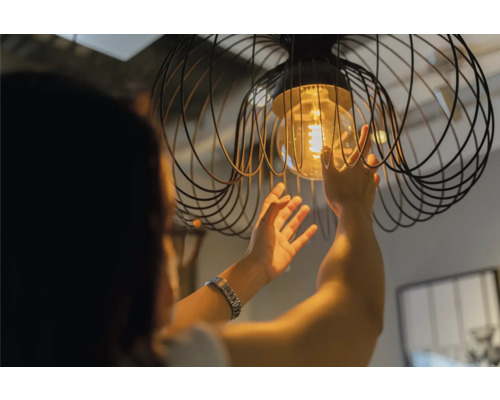 Eine Person wechselt eine Glühbirne in einer Pendelleuchte mit Drahtkäfig.