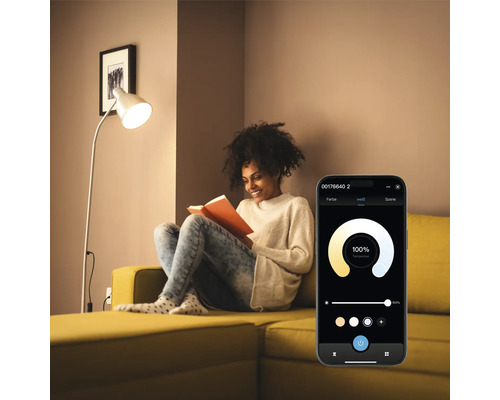 Frau liest auf einem gelben Sofa unter einer Stehlampe mit Lichtsteuerung per Smartphone App.