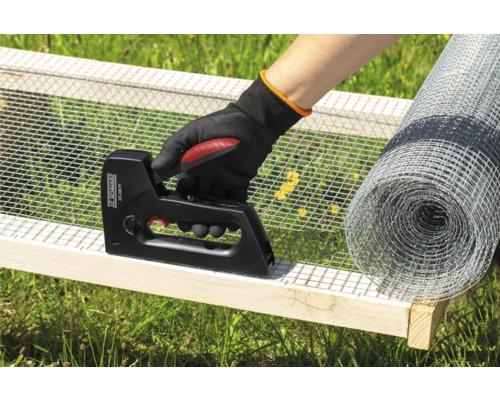 Eine Person befestigt Drahtgeflecht mit einem Tacker an einem Holzrahmen, mit einer Rolle Drahtgeflecht im Hintergrund.