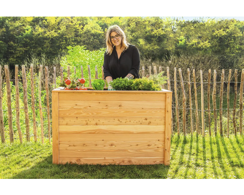 Frau steht hinter einem hölzernen Hochbeet mit verschiedenen Gemüsesorten im Garten