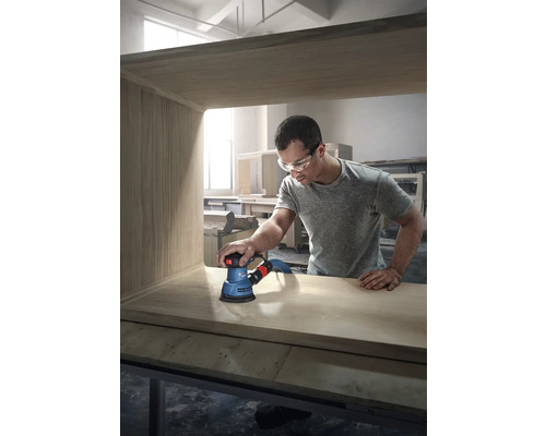 Ein Mann schleift ein Holzmöbelstück mit einem Exzenterschleifer der Marke Bosch.