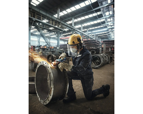Ein Arbeiter schleift mit einer Schleifmaschine ein Rohr in einer Fabrikhalle, dabei trägt er Schutzkleidung.