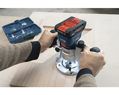 Eine Person benutzt eine Bosch Oberfräse auf einem Holzbrett. Ein Fräserset befindet sich in der Nähe.