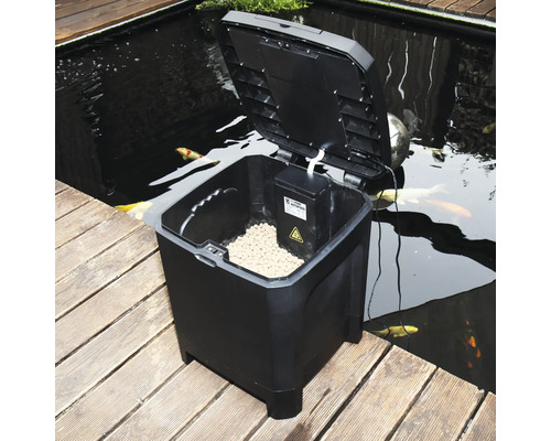 Automatische Fischfuttermaschine mit geöffnetem Deckel neben einem Teich