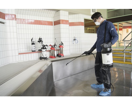 Anwendung von Sprühgeräten in einem gewerblichen Raum mit Fliesenwänden durch eine Person mit Schutzbekleidung.