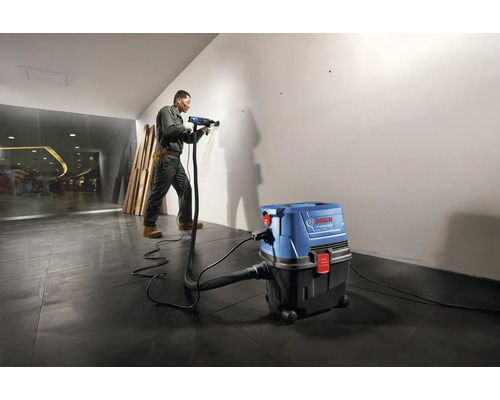 Ein Handwerker bohrt mit einer Bohrmaschine in eine Wand, während ein Industriesauger Staub absaugt.