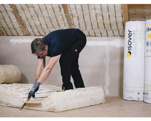 Ein Mann schneidet Dämmwolle im Dachgeschoss zu, wobei Rollen von Isover Dämmmaterial bereitstehen.