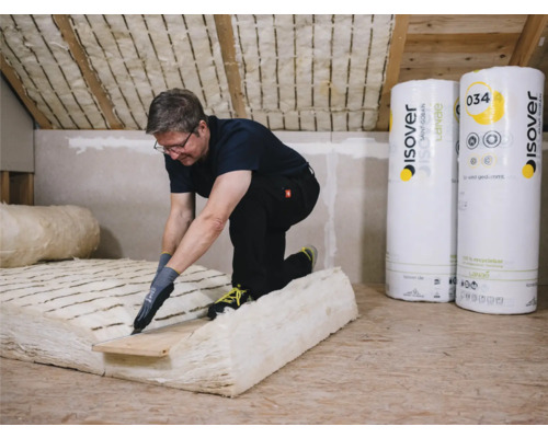 Mann schneidet Dämmmaterial auf einem Dachboden, Rollen des Isover Dämmmaterials im Hintergrund