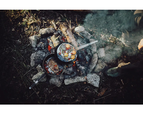 Zwei Bratpfannen mit Speisen auf einem Lagerfeuer, umgeben von Steinen.