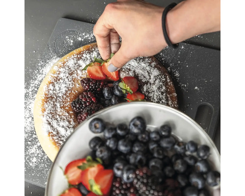 Eine Hand dekoriert einen Kuchen mit Puderzucker, Erdbeeren, Blaubeeren und Brombeeren.