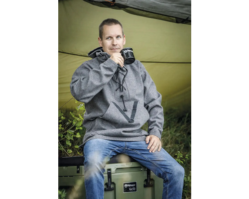 Ein Mann sitzt auf einer Kühlbox und trinkt aus einer Tasse. Er trägt einen grauen Wollpullover.