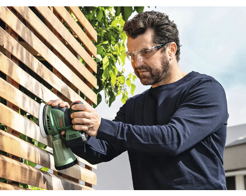 Mann schleift Holzzaun mit einem Akku-Exzenterschleifer der Marke Bosch und trägt dabei eine Schutzbrille.