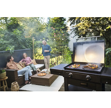 Szene mit Grill, auf dem Essen zubereitet wird, und Personen, die sich auf einer Terrasse unterhalten.