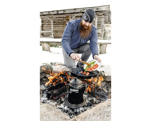 Mann kocht Gemüse in einer Bratpfanne über einem Lagerfeuer im Freien mit einer Kaffeekanne nebenan