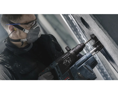 Ein Mann bohrt mit einem Bohrhammer ein Loch in eine Betonwand. Er trägt dabei eine Schutzmaske, eine Schutzbrille und Ohrstöpsel.