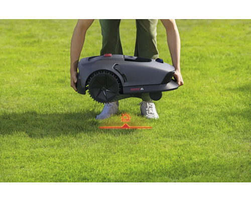 Eine Person trägt einen Mähroboter über einen Rasen.
