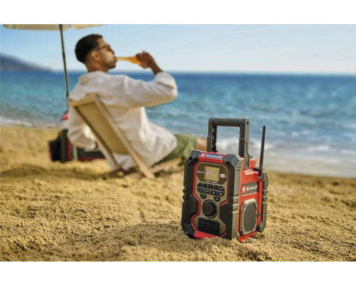 Ein Einhell Baustellenradio am Strand mit einer Person im Hintergrund, die ein Getränk genießt.