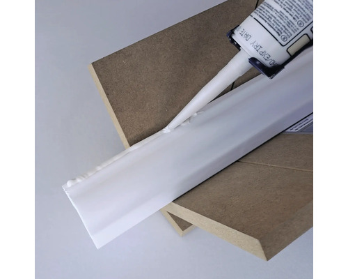 Eine Kartusche Dichtstoff wird auf eine weiße Zierleiste aufgetragen, die auf zwei Holzbrettern liegt.