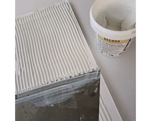 Styroporkleber wird mit einem Spachtel auf eine Platte aufgetragen, im Hintergrund ein Eimer Decosa Kleber