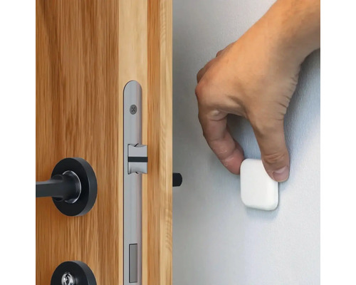 Eine Hand befestigt einen magnetischen Türstopper an einer Wand neben einer Holztür mit einem silberfarbenen Türschloss.