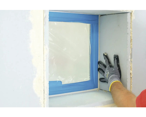 Ein Fensterbereich ist mit Folie und Klebeband abgedeckt, während eine Person mit Handschuhen die Wand berührt