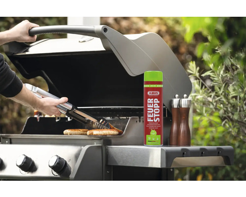 Person grillt Würstchen mit einer Zange auf einem Grill, daneben steht ein Feuerlöschspray.