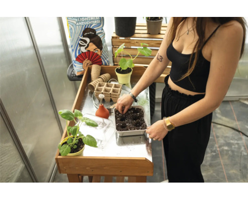 Eine Frau befüllt eine Anzuchtschale mit Erde an einem Tisch im Gewächshaus.